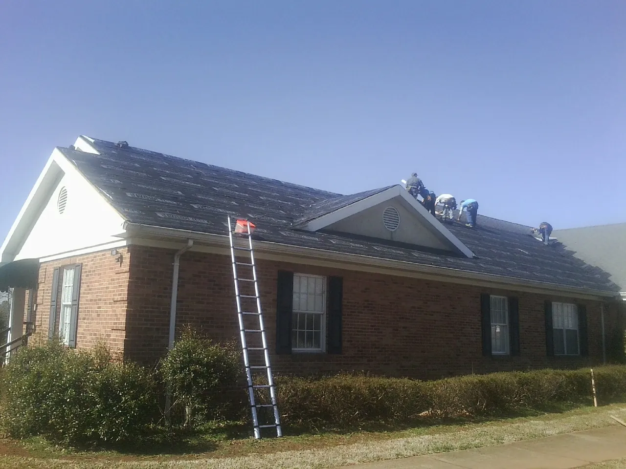 Crew tearing shingles off a brick ranch roof with a ladder set — Emergency Roof Repair work by our crew in Eatonton