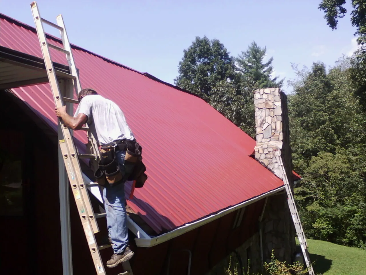 Roofer on a ladder beside a red metal roof and stone chimney — Soffit & Fascia Repair by Roofing Brothers in Eatonton